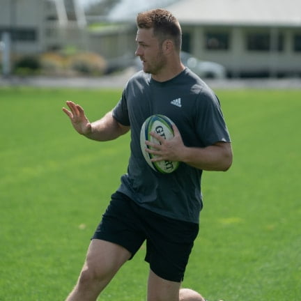 sam cane professional rugby player on a field holding a rugby ball and fending to his right.