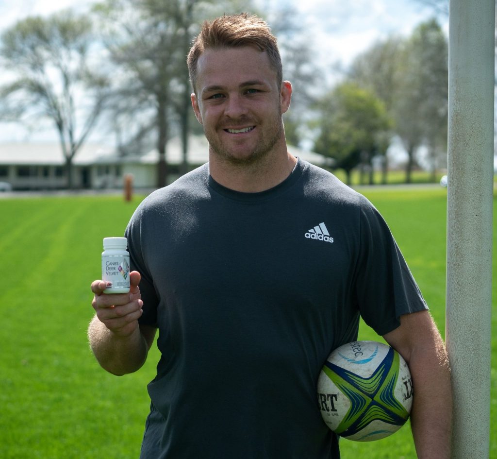 sam cane standing next to a goal post holding a rugby ball and canes deer velvet capsules.