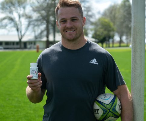 sam cane standing next to a goal post holding a rugby ball and canes deer velvet capsules.